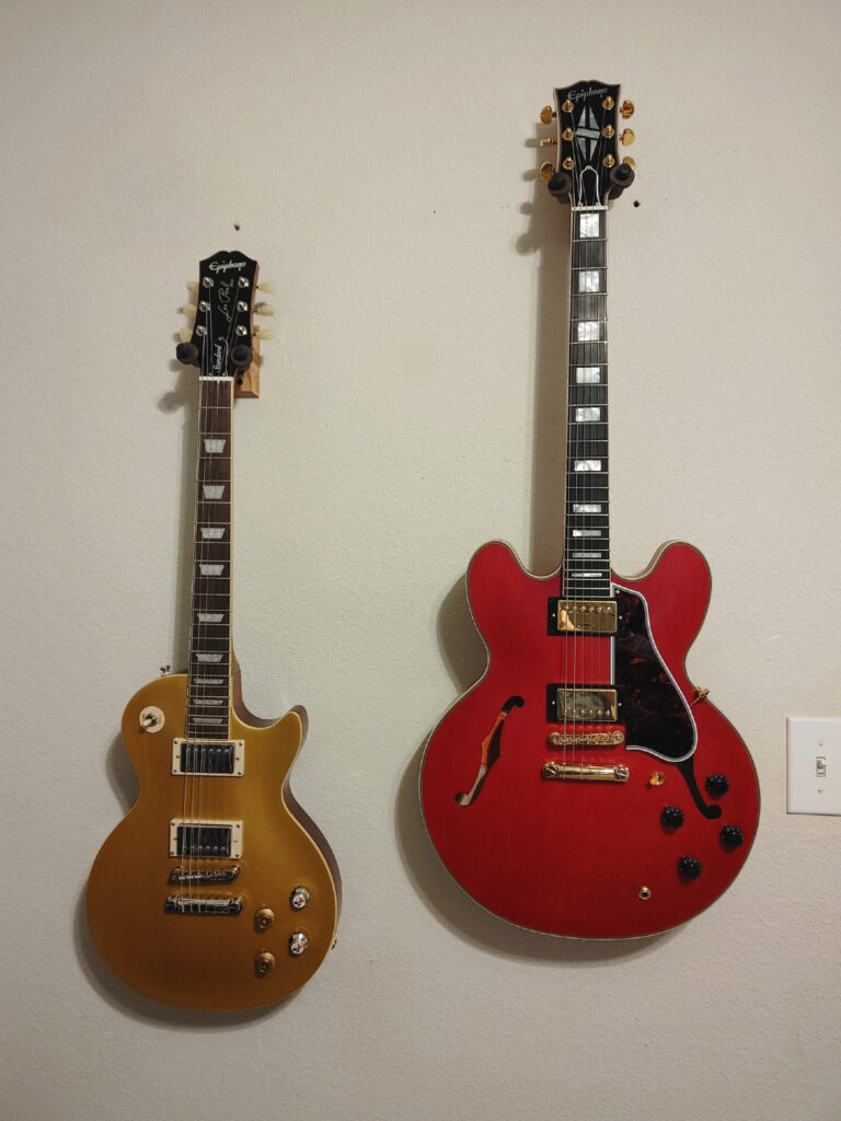 Two guitars are hanging on a wall. On the left is an Epiphone Standard 50's Les Paul. On the right is an Epiphone Inspired by Gibson Custom ES-355.