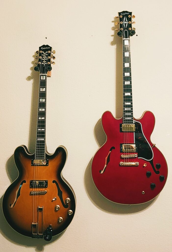 Two guitars are hanging on a wall. On the left is an Epiphone 1962 50th Anniversary Sheraton. On the right is an Epiphone Inspired by Gibson Custom ES-355.